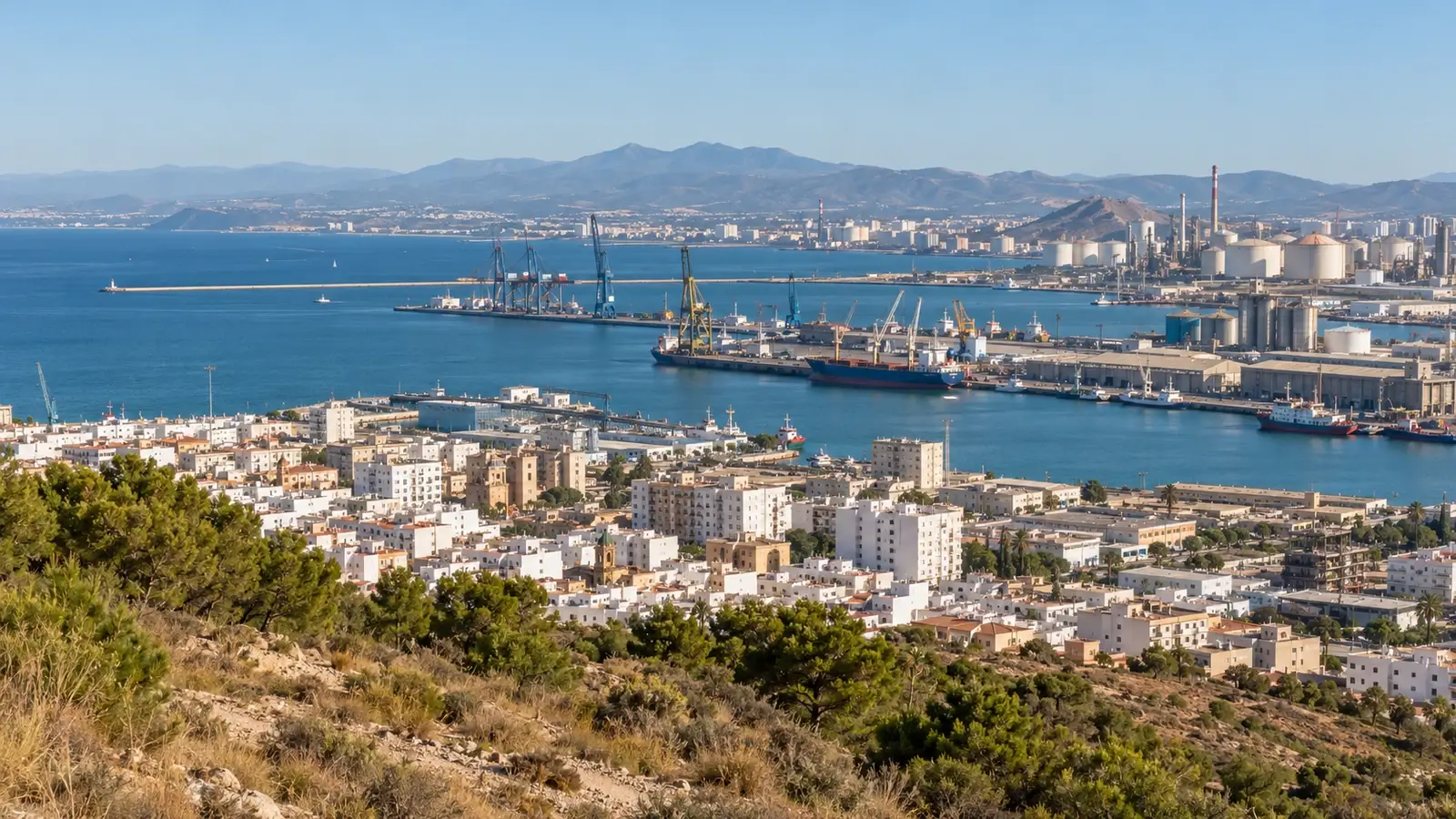 Paisaje costero urbano e industrial con puerto, viviendas y mar bajo un cielo claro